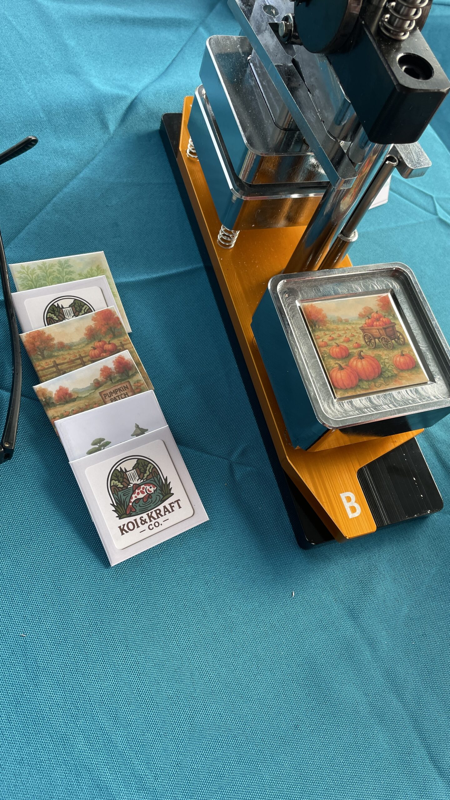 A metal press machine sits on a teal table, surrounded by illustrated cards depicting autumn scenes, showing a crafting setup for making items.