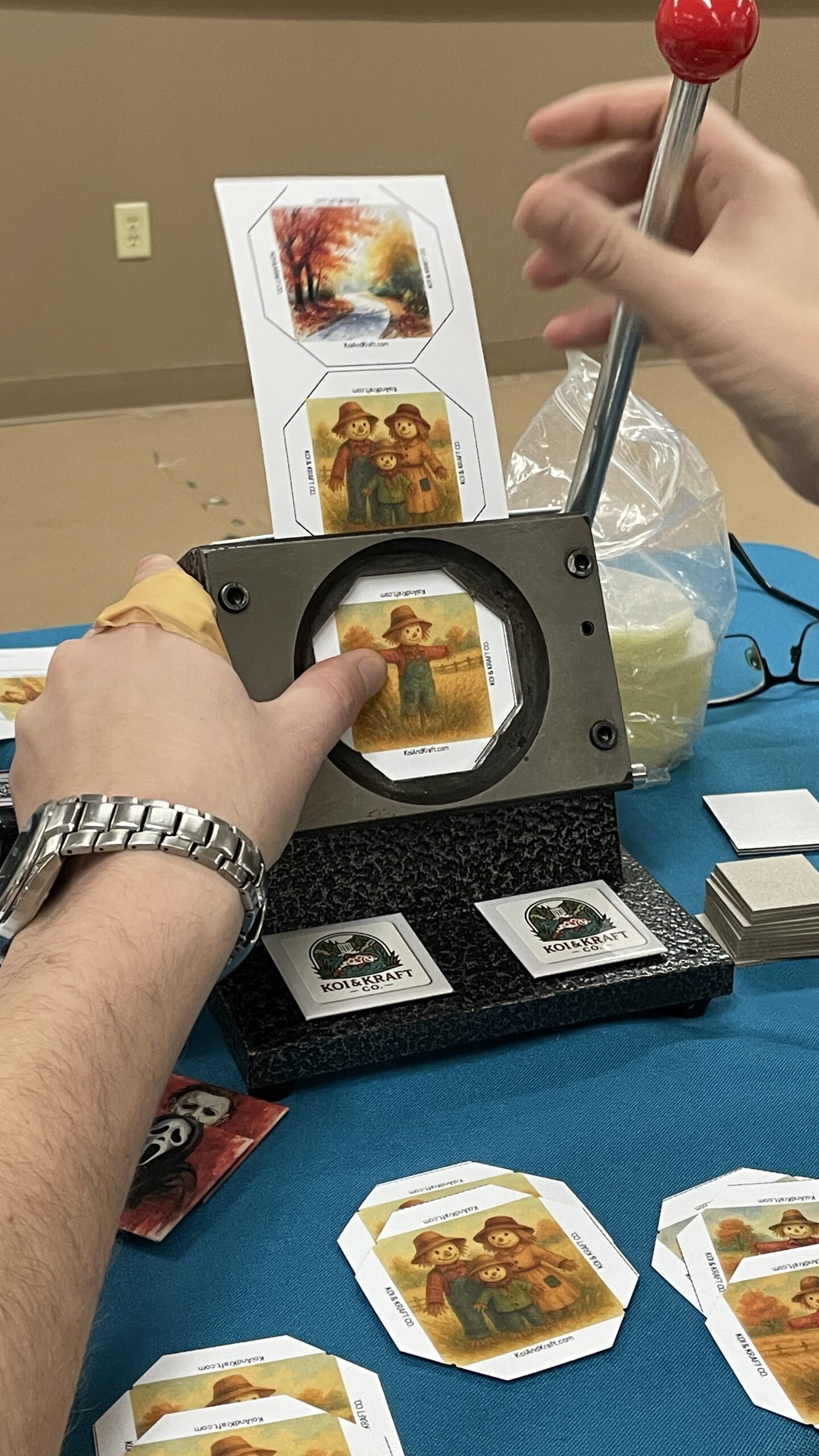 A hand operates a punch machine, cutting out octagonal images of scarecrows and autumn scenes from cardstock. Nearby, additional pre-cut images are arranged.