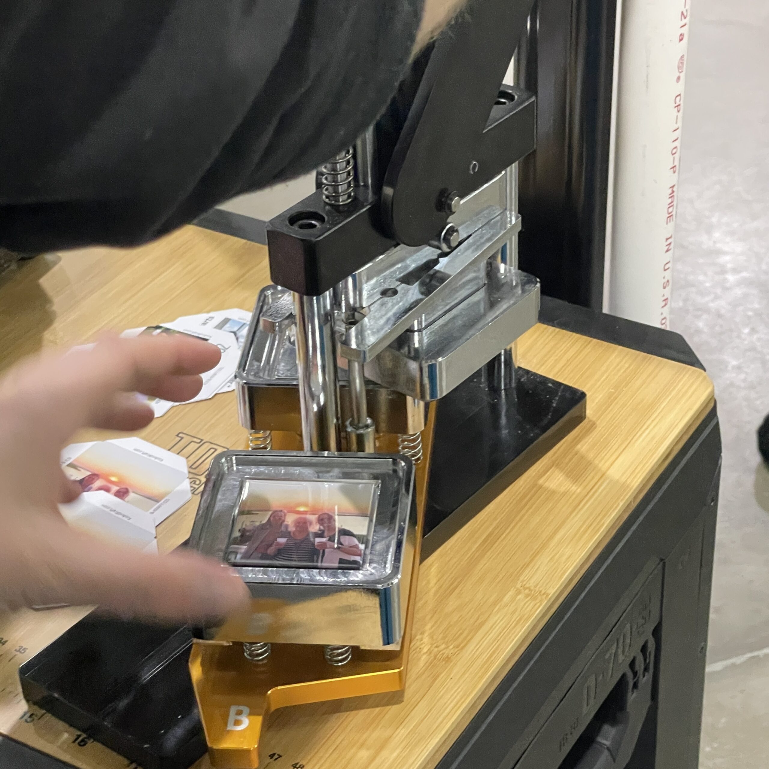 A person operates a photo press, preparing to create a square photo from small printed images. Surrounding materials suggest a crafting or event context.