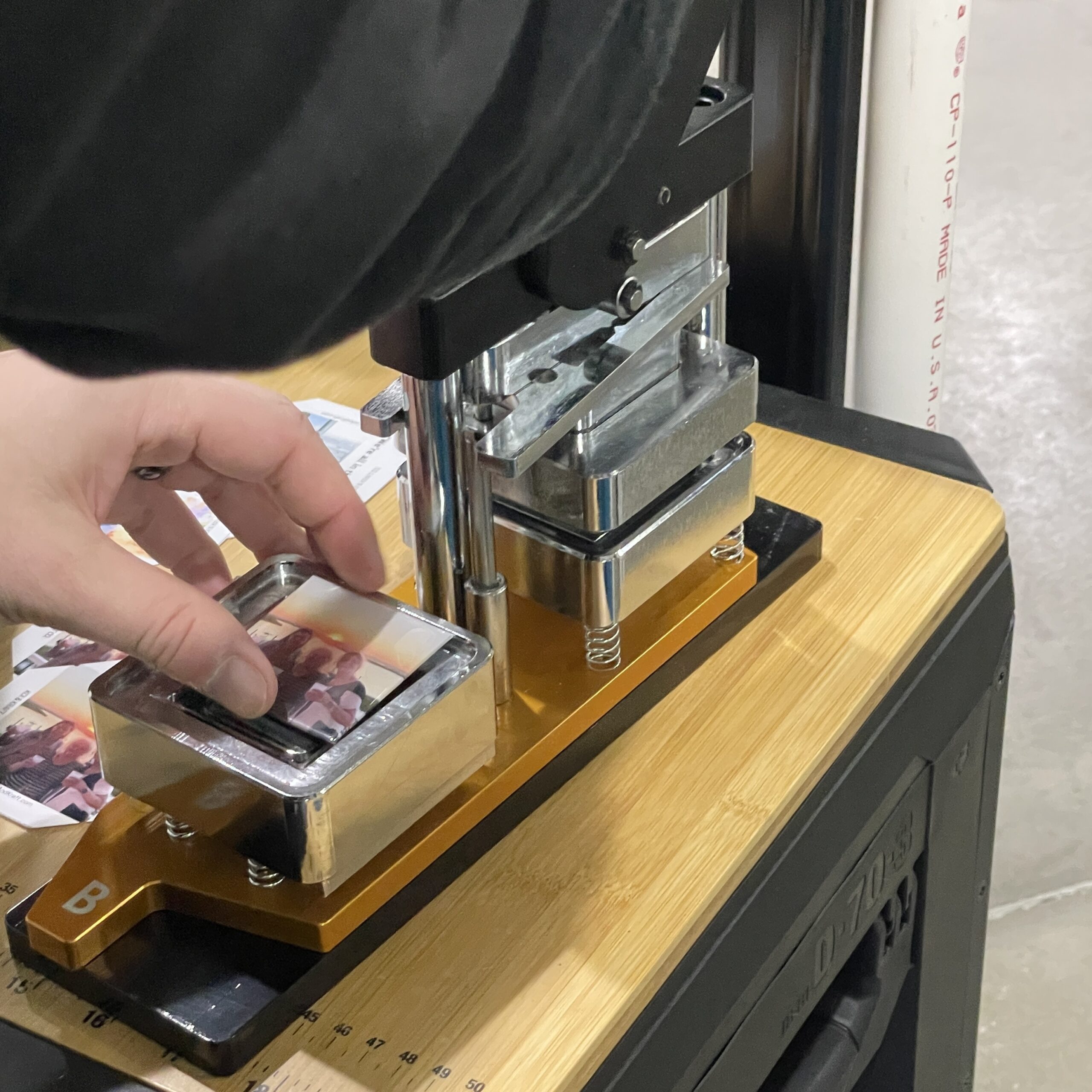 A person presses a photo into a metal mold using a handheld press. Nearby, multiple printed photos are visible, indicating a photo processing task.
